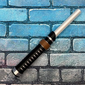 May include: A lightsaber prop with a silver blade and a black and brown handle. The handle has a diamond pattern and silver accents. The prop is set against a blue and gray brick wall.