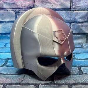 May include: A silver-colored helmet with a geometric design, featuring a visor and a crest with a symbol. The helmet is likely for cosplay or costume use, with a futuristic or sci-fi aesthetic.