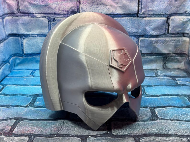 May include: A silver-colored helmet with a geometric design, featuring a visor and a crest with a symbol. The helmet is likely for cosplay or costume use, with a futuristic or sci-fi aesthetic.