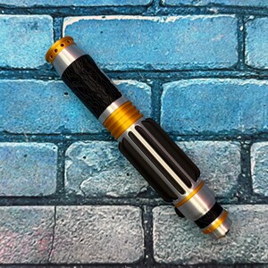 May include: A detailed lightsaber prop with a black, silver, and gold color scheme. The hilt features a textured black grip, silver accents, and gold rings. The top has a gold emitter with small holes. The lightsaber is set against a blue brick background.