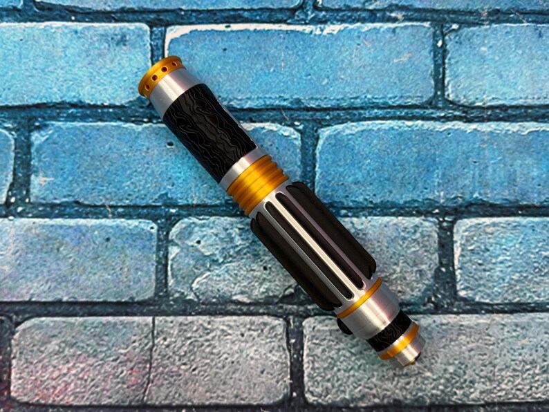 May include: A detailed lightsaber prop with a black, silver, and gold color scheme. The hilt features a textured black grip, silver accents, and gold rings. The top has a gold emitter with small holes. The lightsaber is set against a blue brick background.