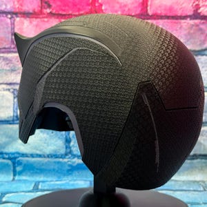 Puede incluir: Un casco negro con una superficie texturizada y una extensi&oacute;n puntiaguda en forma de oreja. El casco se exhibe sobre un soporte negro contra un fondo de pared de ladrillos de colores. El dise&ntilde;o del casco presenta formas geom&eacute;tricas.
