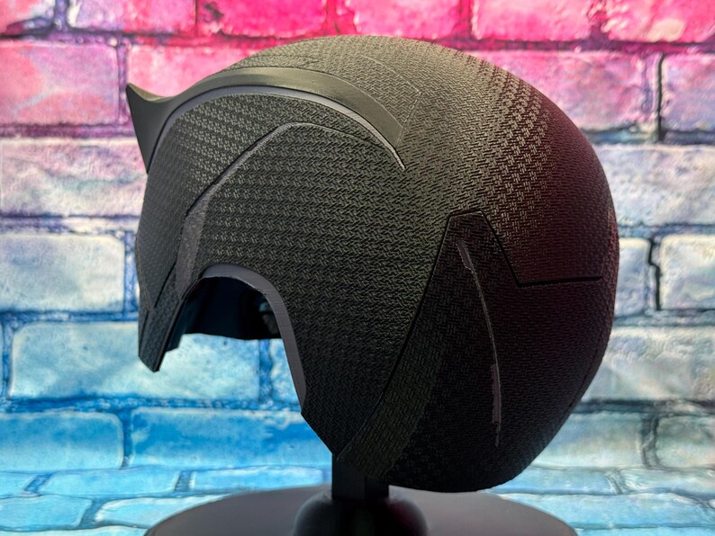 Puede incluir: Un casco negro con una superficie texturizada y una extensi&oacute;n puntiaguda en forma de oreja. El casco se exhibe sobre un soporte negro contra un fondo de pared de ladrillos de colores. El dise&ntilde;o del casco presenta formas geom&eacute;tricas.