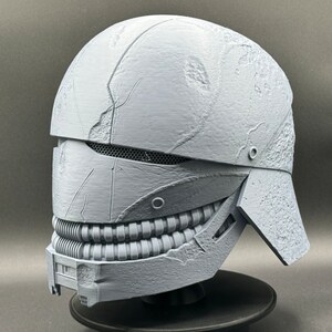 May include: A 3D printed helmet with a textured, weathered surface. The helmet has a visor with a mesh grille and a lower jaw piece with a row of teeth.