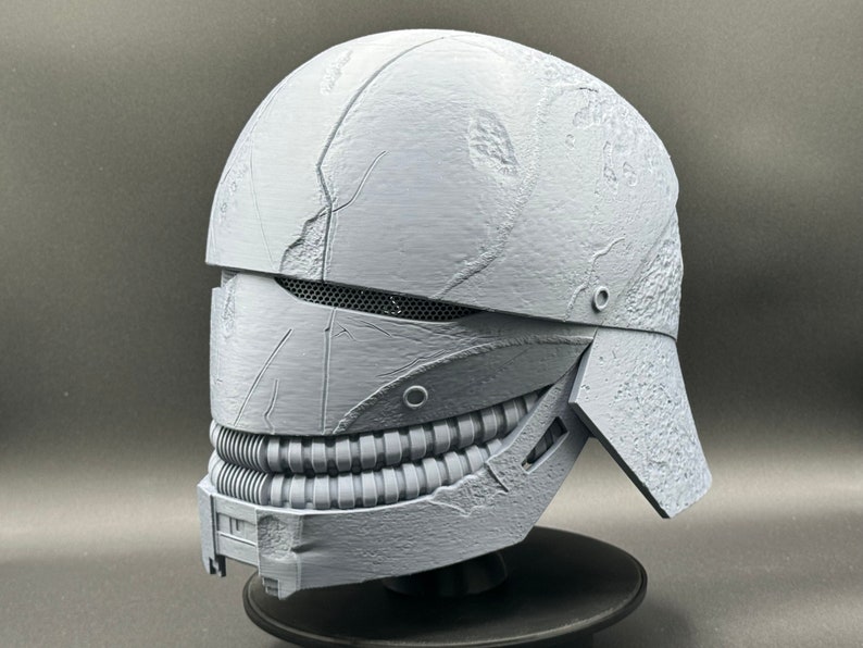 May include: A 3D printed helmet with a textured, weathered surface. The helmet has a visor with a mesh grille and a lower jaw piece with a row of teeth.