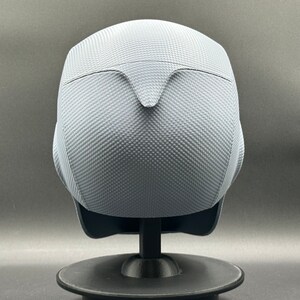 Może przedstawiać: Szary kask z fakturowaną powierzchnią. Kask stoi na czarnym stojaku.