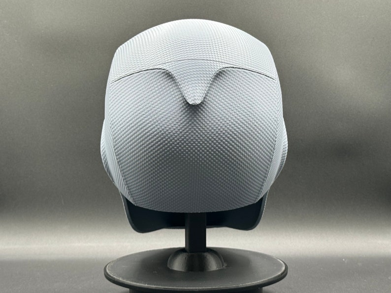 Może przedstawiać: Szary kask z fakturowaną powierzchnią. Kask stoi na czarnym stojaku.