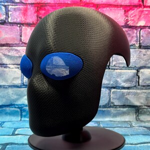 May include: A black full-head mask with a textured, carbon-fiber-like surface finish. The mask features two prominent, oval-shaped blue eye lenses with a fine mesh grid pattern, providing a distinct contrast against the dark, matte-textured material.