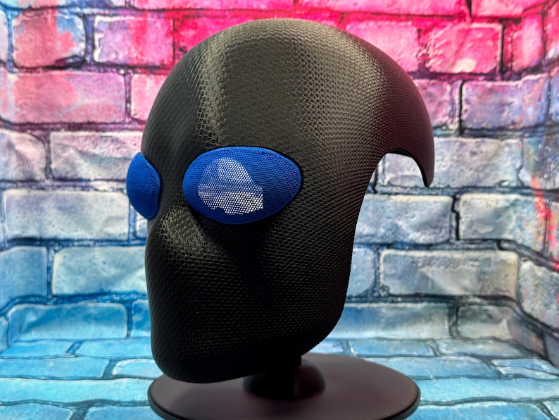 May include: A black full-head mask with a textured, carbon-fiber-like surface finish. The mask features two prominent, oval-shaped blue eye lenses with a fine mesh grid pattern, providing a distinct contrast against the dark, matte-textured material.