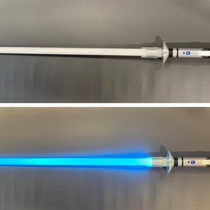 Puede incluir: Un sable de luz plateado y blanco, con una larga hoja cil&iacute;ndrica. La hoja se ilumina con una luz azul brillante en la segunda imagen. El mango presenta detalles plateados y negros, con una guarda circular. Un juguete futurista.