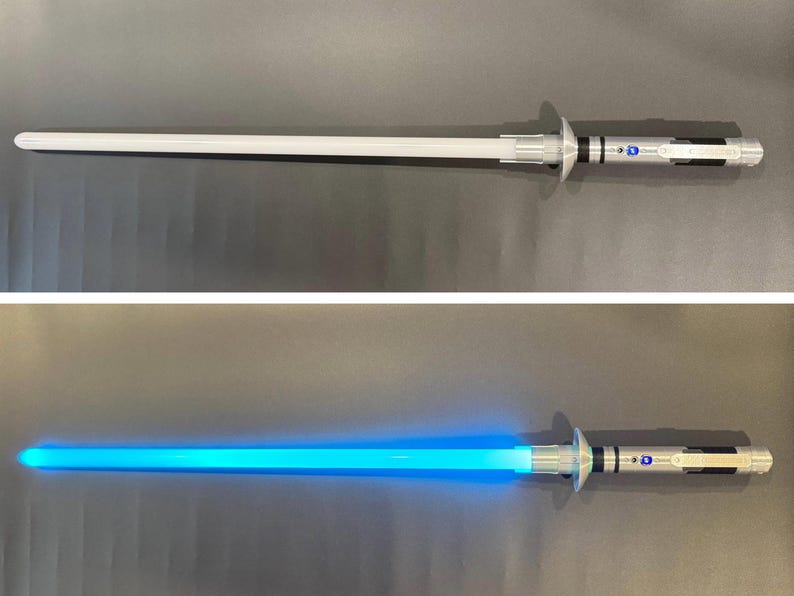 Puede incluir: Un sable de luz plateado y blanco, con una larga hoja cil&iacute;ndrica. La hoja se ilumina con una luz azul brillante en la segunda imagen. El mango presenta detalles plateados y negros, con una guarda circular. Un juguete futurista.