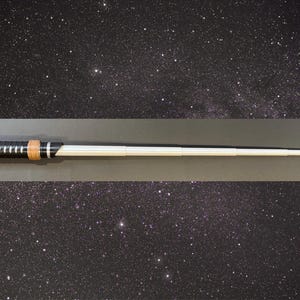 May include: A black and silver lightsaber prop with a brown grip and a white blade. The lightsaber is set against a starry background, suggesting a space or sci-fi theme. The prop is a replica of a lightsaber.