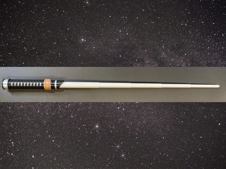 May include: A black and silver lightsaber prop with a brown grip and a white blade. The lightsaber is set against a starry background, suggesting a space or sci-fi theme. The prop is a replica of a lightsaber.