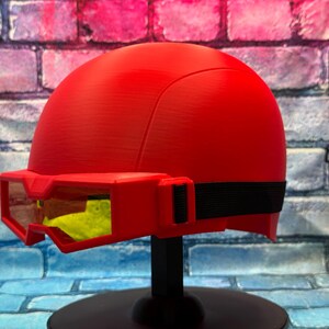 May include: A bright red helmet featuring a smooth, rounded dome design with an integrated red visor frame. The visor contains yellow-tinted lenses and is secured to the sides of the helmet with a black elastic strap, creating a cohesive look.
