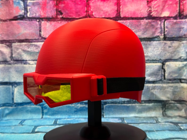 May include: A bright red helmet featuring a smooth, rounded dome design with an integrated red visor frame. The visor contains yellow-tinted lenses and is secured to the sides of the helmet with a black elastic strap, creating a cohesive look.