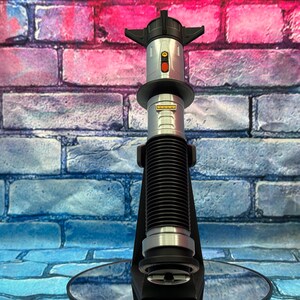 K&ouml;nnte beinhalten: Ein silbernes und schwarzes Lichtschwert-Requisit mit einem geriffelten schwarzen Griff und einem roten Knopf. Das Lichtschwert ist auf einem schwarzen St&auml;nder mit kreisf&ouml;rmiger Basis montiert. Der Hintergrund ist eine Backsteinmauer mit blauen, rosa und violetten Farbt&ouml;nen.