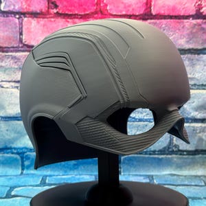 May include: A gray helmet with a textured surface and angular design, featuring a visor opening and a chin strap. The helmet is displayed on a black stand against a brick wall background.