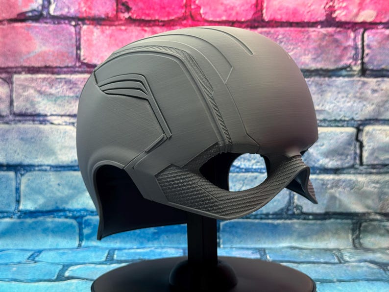 May include: A gray helmet with a textured surface and angular design, featuring a visor opening and a chin strap. The helmet is displayed on a black stand against a brick wall background.
