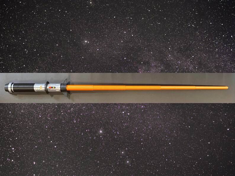 K&ouml;nnte beinhalten: Ein Lichtschwert-Requisite mit einem schwarz-silbernen Griff und einer orangefarbenen Klinge. Der Griff hat eine strukturierte Oberfl&auml;che und silberne Akzente. Die Klinge ist leuchtend orange und verj&uuml;ngt sich zu einer Spitze. Das Lichtschwert wird vor einem Sternenhintergrund pr&auml;sentiert.