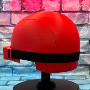 May include: A smooth, matte red helmet featuring a rounded dome shape and a black elastic strap that wraps around the sides. The helmet includes a small red rectangular attachment on the left side where the black strap connects to the main shell.
