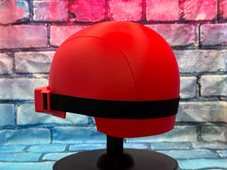 May include: A smooth, matte red helmet featuring a rounded dome shape and a black elastic strap that wraps around the sides. The helmet includes a small red rectangular attachment on the left side where the black strap connects to the main shell.