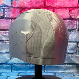 May include: A silver-colored helmet with a smooth, curved design. The helmet has a segmented appearance with a textured surface. The right side of the helmet is a reddish-pink color. The helmet is displayed on a black stand.