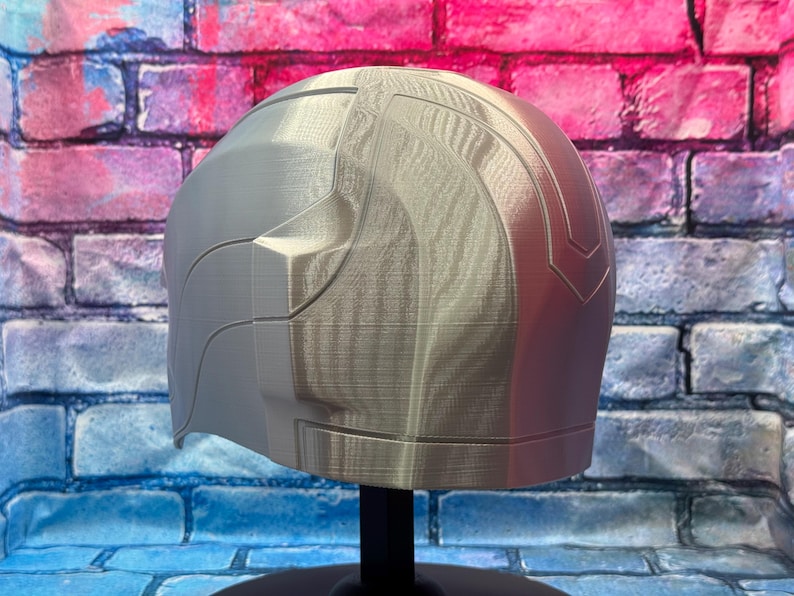 May include: A silver-colored helmet with a smooth, curved design. The helmet has a segmented appearance with a textured surface. The right side of the helmet is a reddish-pink color. The helmet is displayed on a black stand.