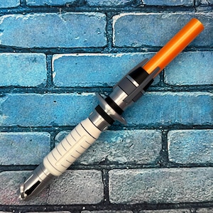 May include: A futuristic-looking lightsaber prop with a white, silver, and black handle and an orange blade. The handle has a ribbed design and is set against a blue brick wall.