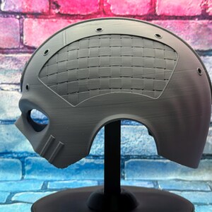 May include: A gray, half-face mask with an eye opening and a woven pattern on the top. The mask has a curved shape and is mounted on a black stand. The background is a brick wall.