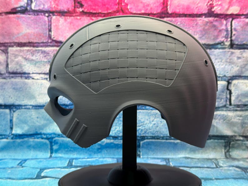 May include: A gray, half-face mask with an eye opening and a woven pattern on the top. The mask has a curved shape and is mounted on a black stand. The background is a brick wall.