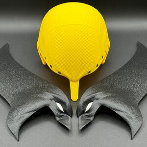 Puede incluir: Un casco amarillo con alas negras a cada lado. El casco tiene una superficie texturizada y varios agujeros peque&ntilde;os. Las alas tambi&eacute;n est&aacute;n texturizadas y tienen una forma de ojo blanca a cada lado.