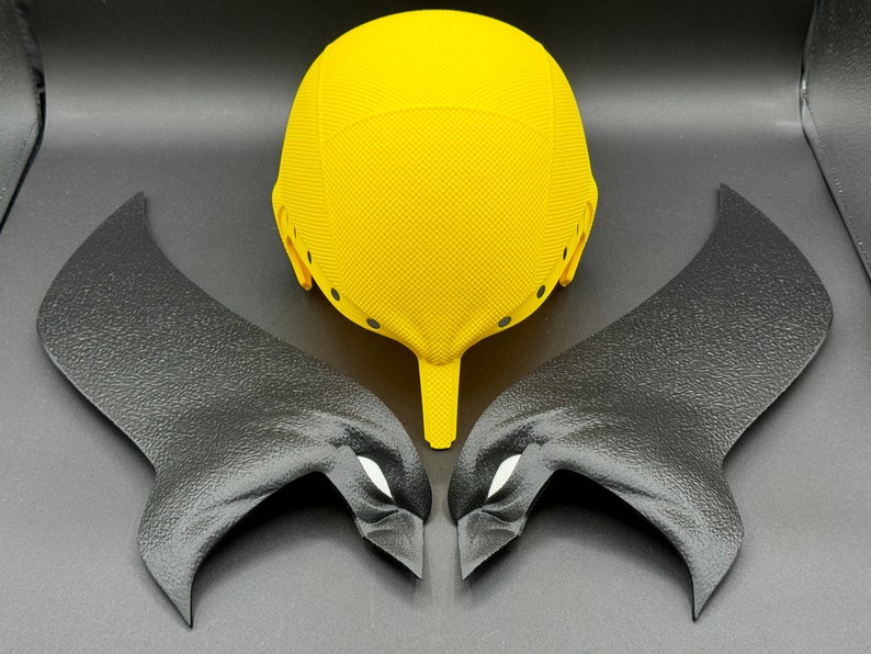 Puede incluir: Un casco amarillo con alas negras a cada lado. El casco tiene una superficie texturizada y varios agujeros peque&ntilde;os. Las alas tambi&eacute;n est&aacute;n texturizadas y tienen una forma de ojo blanca a cada lado.