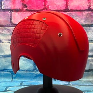 May include: A bright red helmet with a textured, woven-like pattern on the left side. The helmet has a smooth, curved design with three small, silver-colored rivets. It is displayed on a black stand against a colorful brick wall.