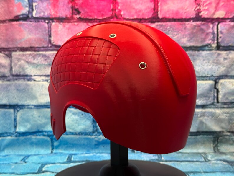 May include: A bright red helmet with a textured, woven-like pattern on the left side. The helmet has a smooth, curved design with three small, silver-colored rivets. It is displayed on a black stand against a colorful brick wall.