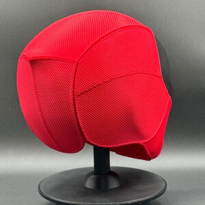 May include: A red and black Deadpool mask with a textured surface. The mask is on a black stand.