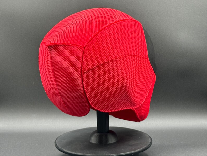 May include: A red and black Deadpool mask with a textured surface. The mask is on a black stand.