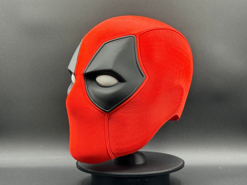 K&ouml;nnte beinhalten: Eine rote und schwarze Deadpool-Maske mit einem wei&szlig;en Augennetz. Die Maske steht auf einem schwarzen St&auml;nder.
