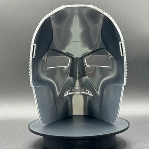 May include: A black plastic mask with a detailed design. The mask has two eye holes and a mouth hole. The mask is on a black stand.