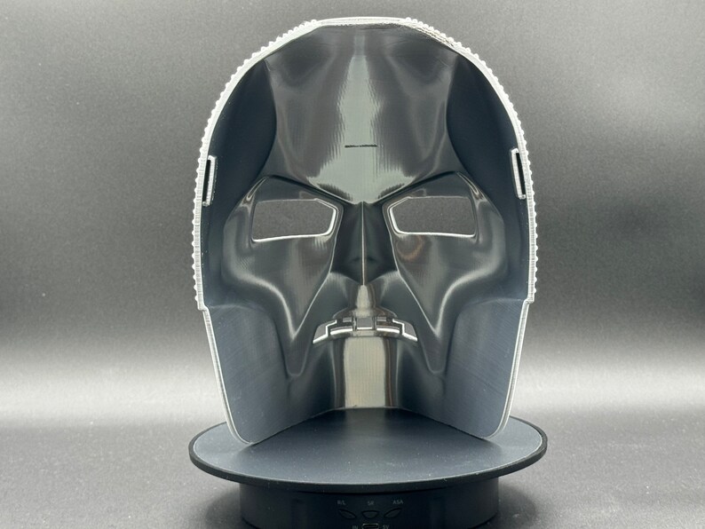 May include: A black plastic mask with a detailed design. The mask has two eye holes and a mouth hole. The mask is on a black stand.
