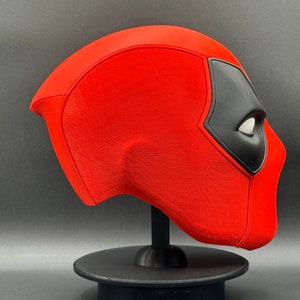 K&ouml;nnte beinhalten: Eine rote und schwarze Deadpool-Maske auf einem schwarzen St&auml;nder. Die Maske besteht aus einem strukturierten Material und hat ein detailliertes Design.