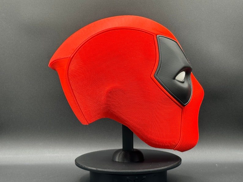 K&ouml;nnte beinhalten: Eine rote und schwarze Deadpool-Maske auf einem schwarzen St&auml;nder. Die Maske besteht aus einem strukturierten Material und hat ein detailliertes Design.