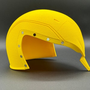 May include: A yellow helmet with a textured surface and multiple silver accents. The helmet is open on one side and has a pointed end.