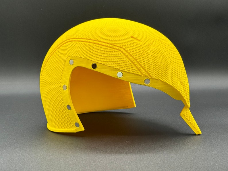 May include: A yellow helmet with a textured surface and multiple silver accents. The helmet is open on one side and has a pointed end.