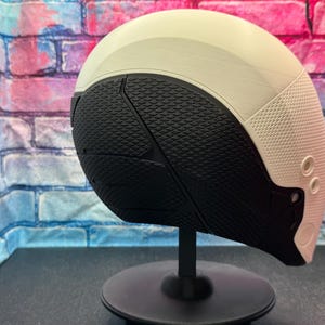May include: A futuristic-looking helmet with a cream-colored top and a textured black lower section. The helmet has a rounded shape and is mounted on a black stand. The design includes small circular details on the side.