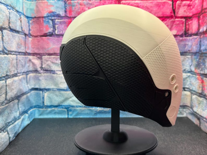 May include: A futuristic-looking helmet with a cream-colored top and a textured black lower section. The helmet has a rounded shape and is mounted on a black stand. The design includes small circular details on the side.