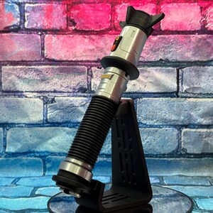 K&ouml;nnte beinhalten: Ein silbernes und schwarzes Lichtschwert-Requisit mit geriffeltem schwarzem Griff und rotem Knopf. Das Lichtschwert wird auf einem schwarzen St&auml;nder vor einem Backsteinmauer-Hintergrund ausgestellt.