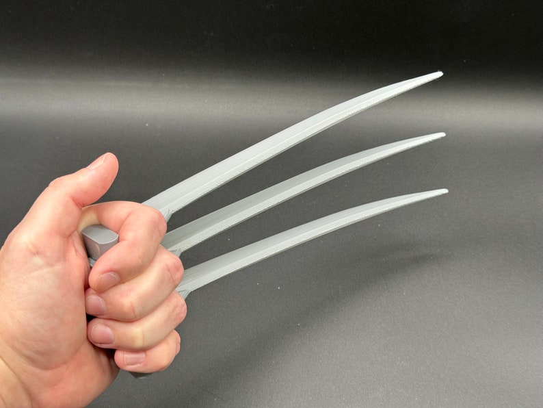 May include: Three silver-colored metal claws, resembling Wolverine's claws, are held in a hand. The claws are sharp and pointed, with a slightly curved shape.