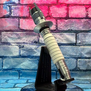 May include: A futuristic lightsaber prop with a black and silver hilt and a white, ribbed grip. The prop is displayed on a black stand against a brick wall backdrop with pink, purple, and blue hues.