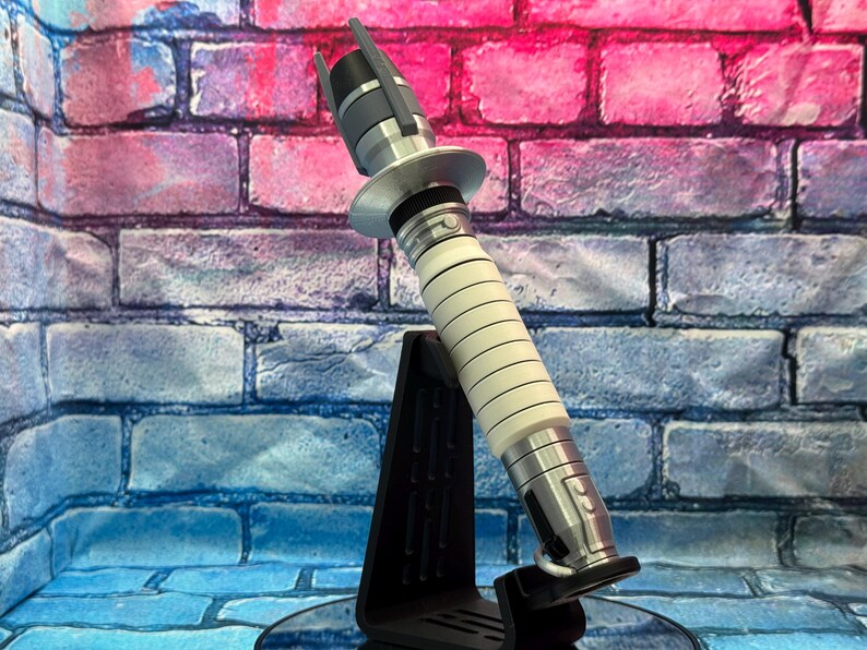 May include: A futuristic lightsaber prop with a black and silver hilt and a white, ribbed grip. The prop is displayed on a black stand against a brick wall backdrop with pink, purple, and blue hues.