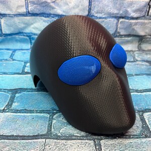May include: A dark brown, textured, full-face mask featuring a smooth, carbon-fiber-like finish. The mask includes two prominent, oval-shaped eye cutouts covered with bright blue, fine-mesh material, creating a distinct contrast against the dark shell.